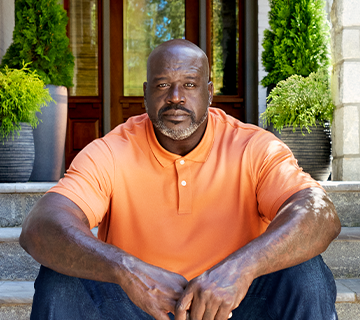 Shaquille O'Neal in an orange shirt and jeans sitting on a front porch